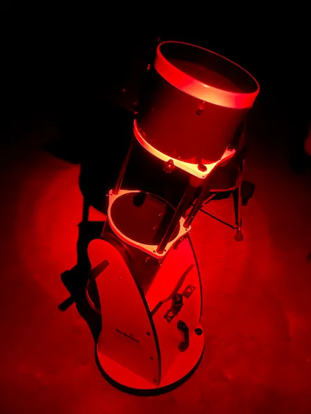 Dobsonian telescope glowing under red light at a star party, photographed from an angle showing the full setup