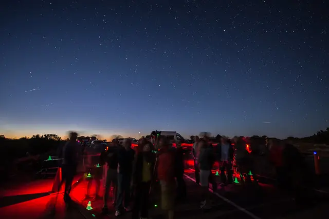 A group of people stargazing under a star-filled sky at sunset, with telescopes and red lights.